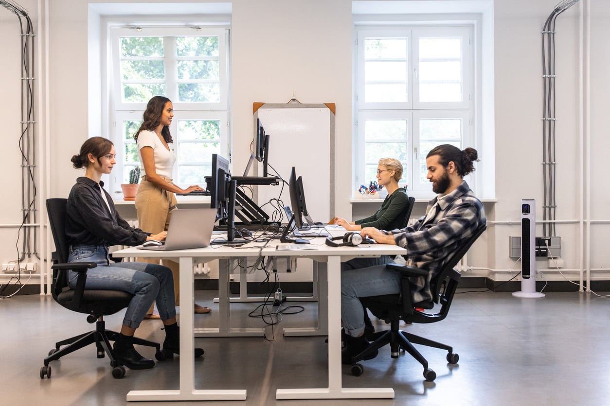 Employees sitting and standing at sit-stand workstations in a shared office environment.